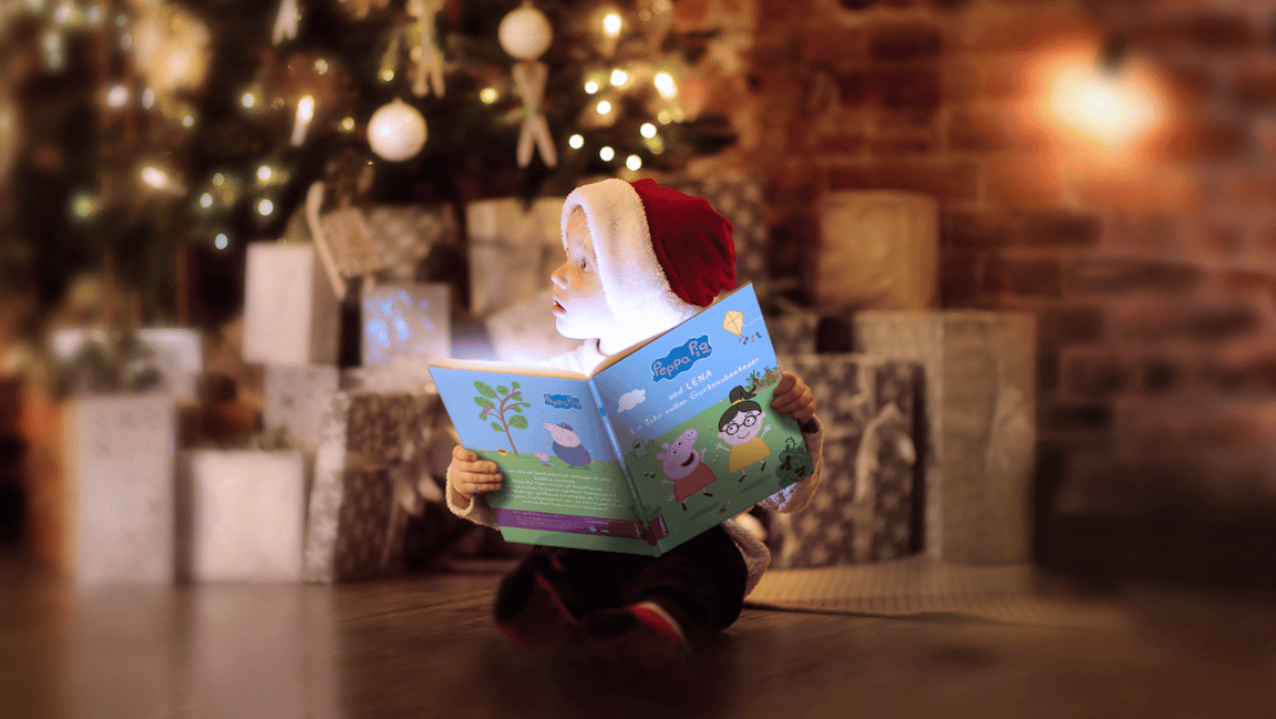 Kleiner Junge sitzt mit dem personalisierten Kinderbuch Peppa Pig im Gartenabenteuer vor dem Tannenbaum mit vielen Geschenken im Hintergrund