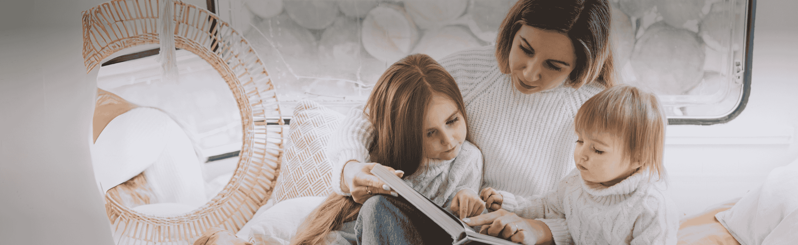 Eine Frau sitzt auf einem Bett in einem gemütlichen Zimmer und liest zwei kleinen Kindern neben ihr eine Geschichte vor, die alle in warme, helle Kleidung gekleidet sind.