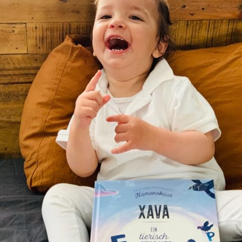 Ein kleines Kind in einem weißen Outfit sitzt auf einem Bett mit senffarbenen Kissen, lächelt und hält ein personalisiertes Buch mit dem Titel Xava - Ein tierisch starker Name.