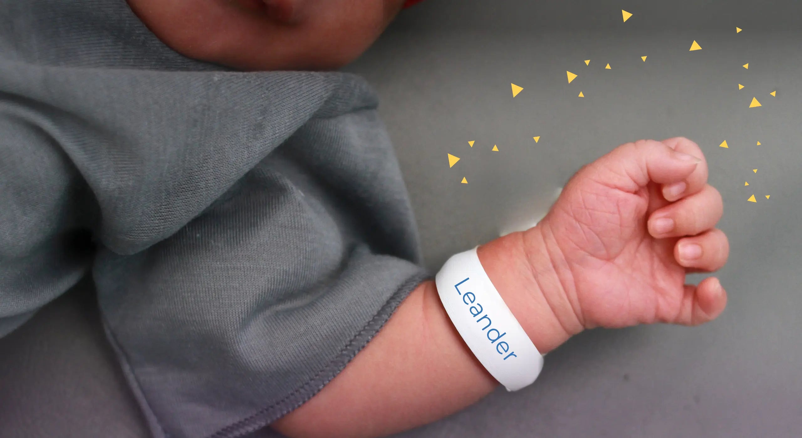 Eine Nahaufnahme des Arms eines Neugeborenen, das ein weißes Namensarmband mit der Aufschrift Leander trägt. Das Baby ist in eine kuschelige graue Decke eingewickelt und auf dem weichen Stoff sind kleine gelbe Dreiecke verstreut.