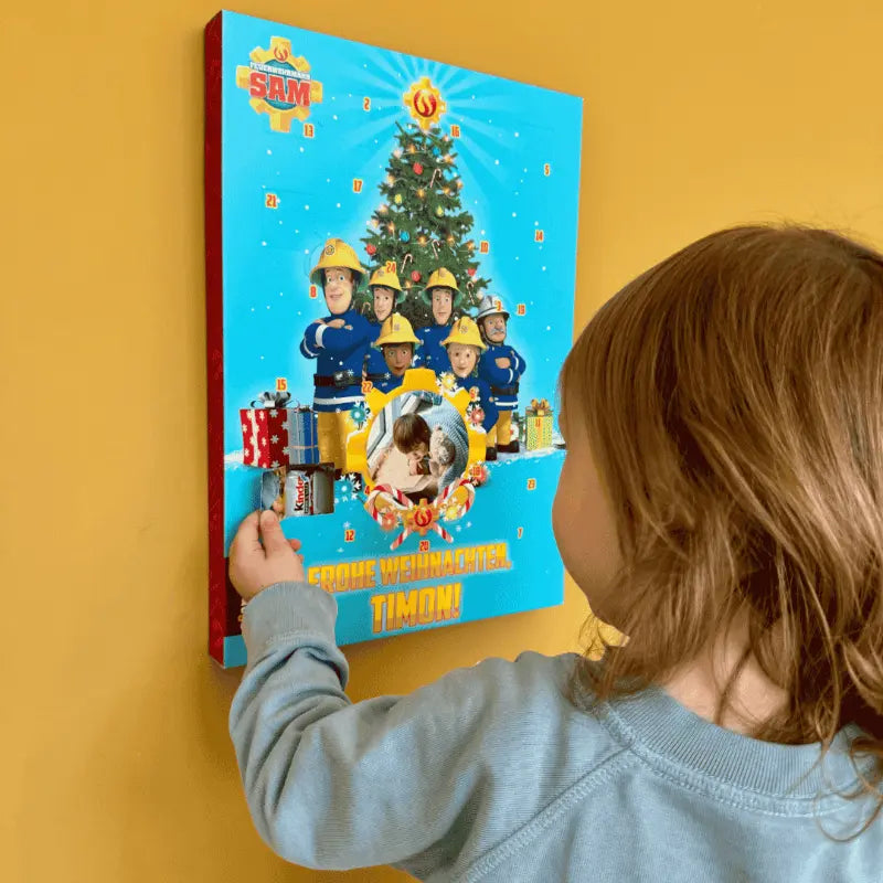 Ein kleines Kind in einem blauen Hemd öffnet eine Tür des Adventskalenders von Framily Feuerwehrmann Sam, der mit einem Weihnachtsbaum, Feuerwehrleuten und Geschenken geschmückt ist und an einer gelben Wand hängt.