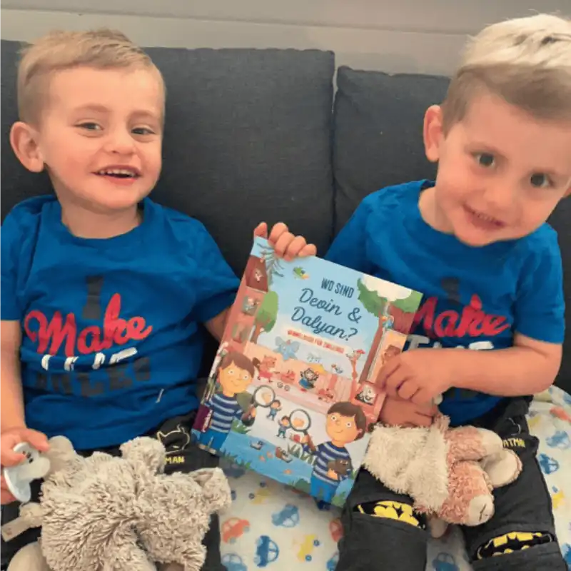 Zwei kleine Jungen mit kurzen Haaren und blauen Hemden sitzen lächelnd auf einem Sofa, jeder hält ein Stofftier und zeigt stolz das Framily-Wimmelbuch für Geschwister: „Wer sind Devin & Daylan?“.